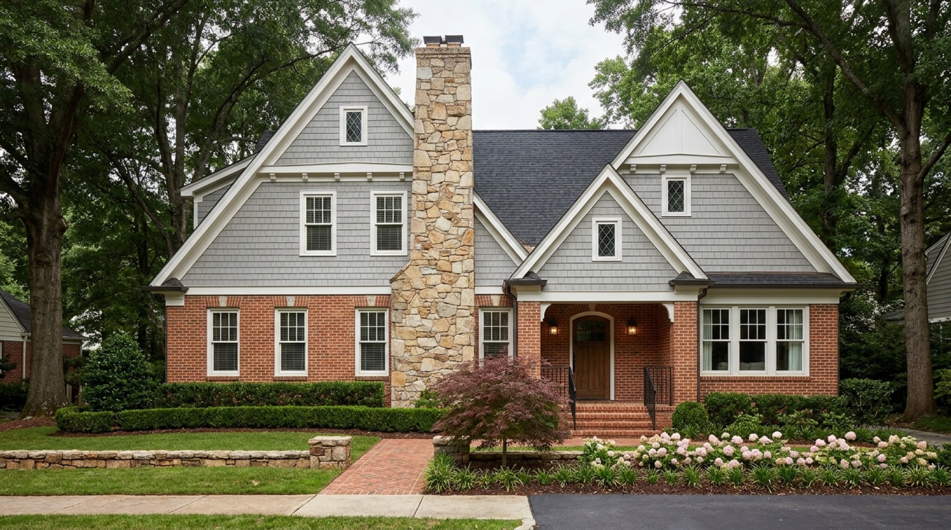 Two-story Tudor Revival home with steep front-facing gables, dove-grey shingle siding, and a hand-laid fieldstone chimney rising through the center of the facade — Druid Hills / Ansley Park vernacular.