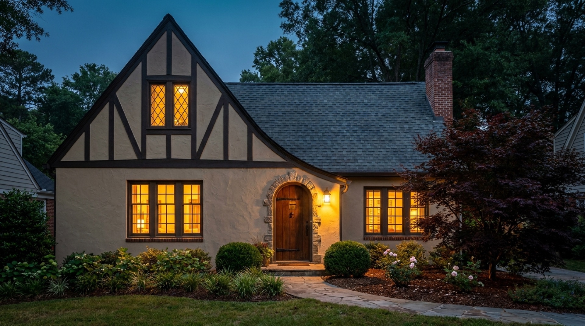 English storybook cottage at deep dusk with every window glowing warm from within, stucco and dark half-timbering framed by a deep-indigo twilight sky.